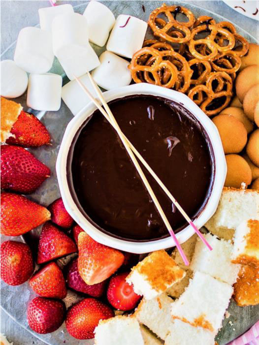 A bowl of how to make orange fused dark chocolate fondue surrounded by strawberries, cubed cake, marshmallows, pretzels, and vanilla wafers. Two skewers are placed in the chocolate.