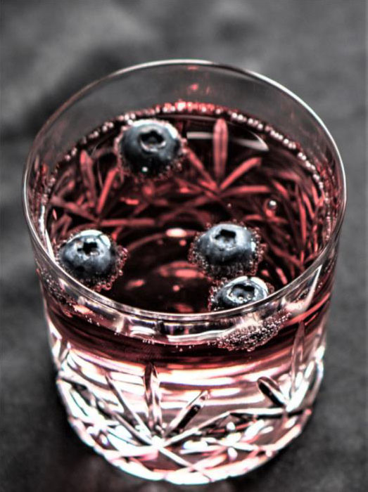 A glass of red liquid garnished with four floating blueberries on a dark background, reminiscent of how to make blueberry lemon-thyme balsamic sparkling shrub.