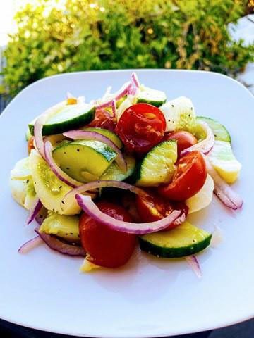 A plate of fresh salad with sliced cucumbers, cherry tomatoes, red onions, and cheese chunks on a white square dish, set against a blurred outdoor background. Learn how to make cucumber summer salad that’s perfect for any picnic or gathering.