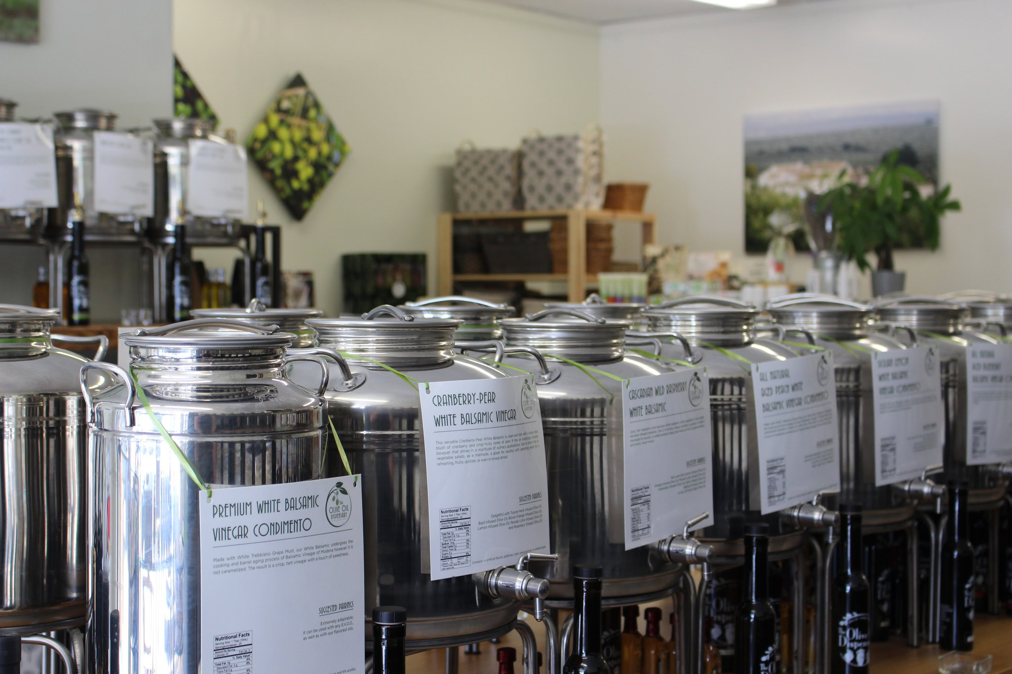 A row of stainless steel containers on a counter in a store for dispensing various types of extra virgin olive oil and vinegar. Shelves with additional products are visible in the background.