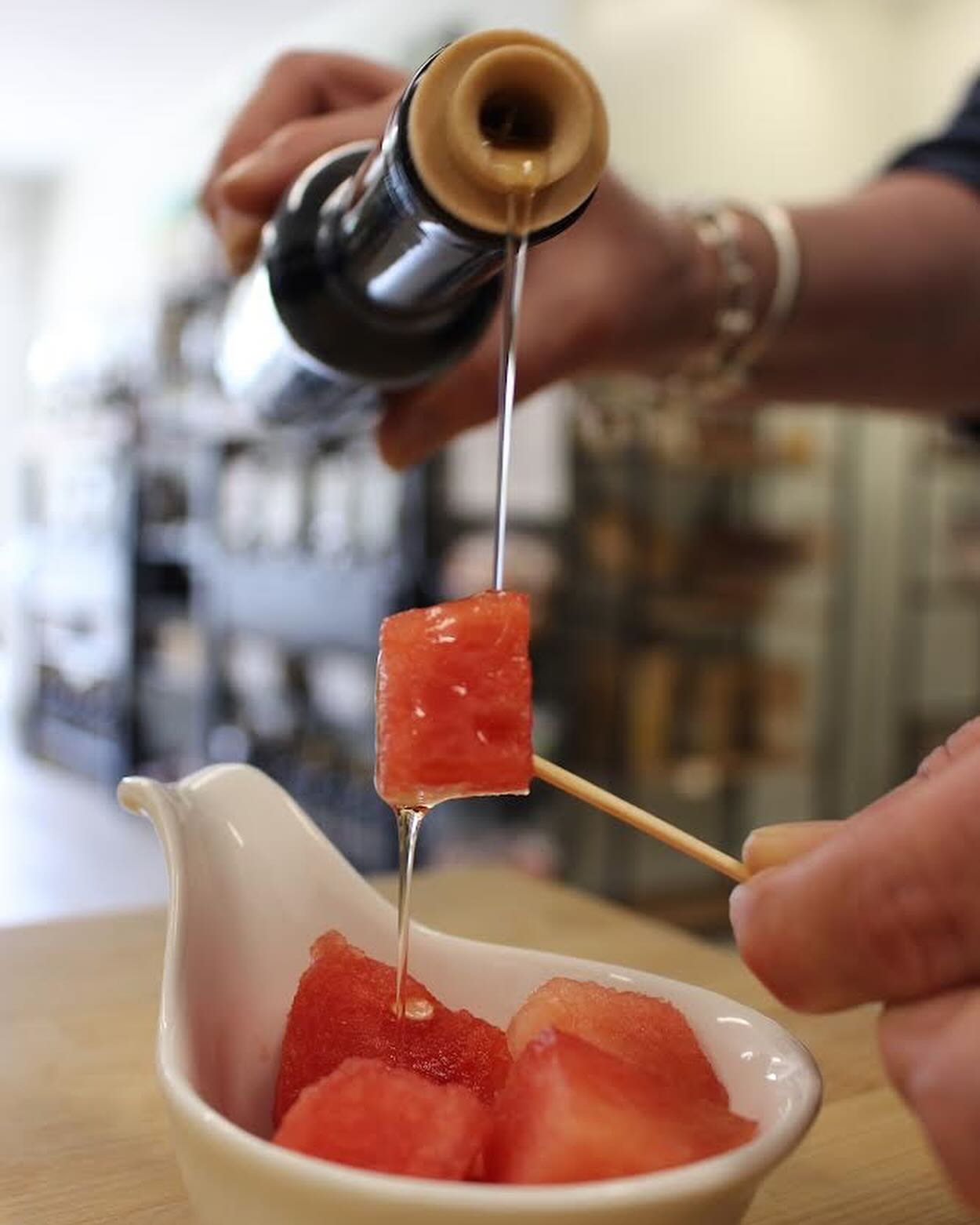 A person pours syrup over a skewer with a watermelon cube, held above a bowl containing more watermelon cubes. A drizzle of extra virgin olive oil enhances the flavors, creating a delightful summer treat.