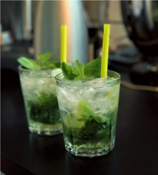 Two glasses of mojitos with yellow straws, garnished with fresh mint leaves and ice, placed on a dark surface. Learn how to make lemongrass-mint mojito for a refreshing twist.