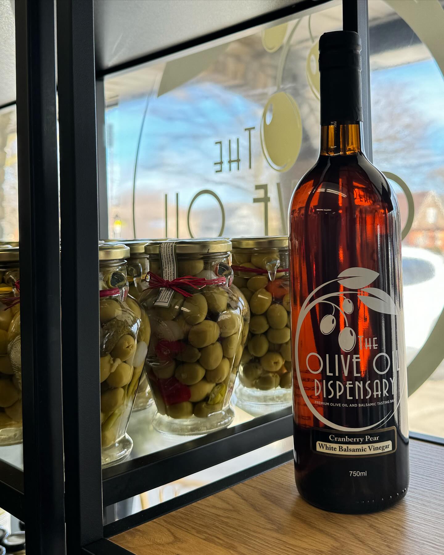 A bottle of cranberry pear white balsamic vinegar, elegantly labeled "The Olive Oil Dispensary," is displayed on a shelf beside jars filled with vibrant green olives.