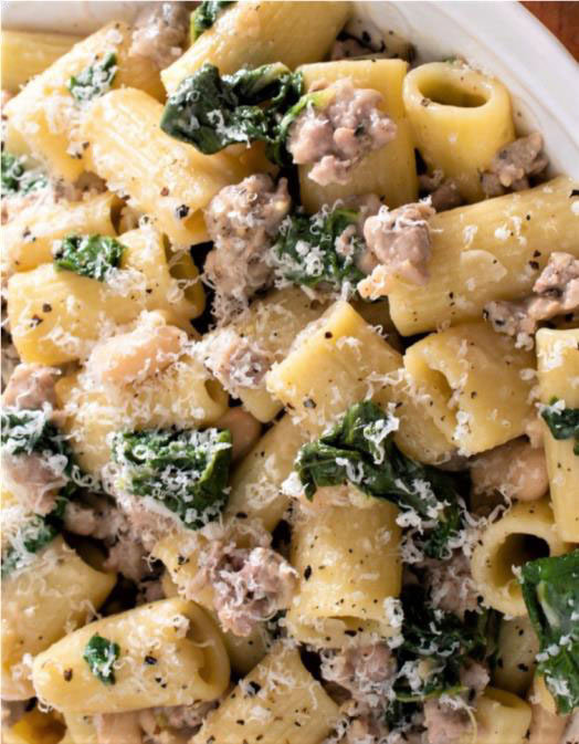 A close-up of a pasta dish featuring rigatoni, sautéed spinach, ground meat, and grated cheese showcases a delicious meal. Learn how to make Rigatoni with Sausage for an irresistible dinner tonight.