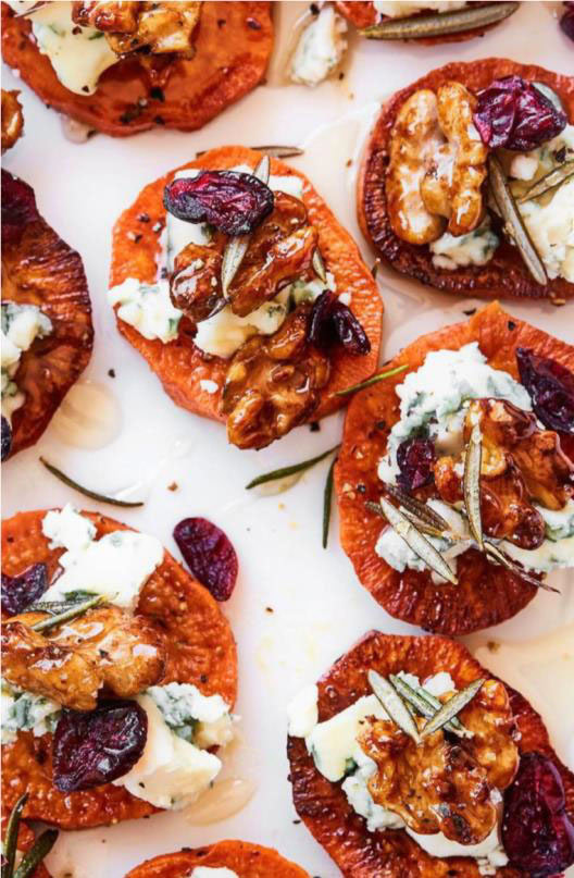 Close-up of sweet potato rounds topped with blue cheese, walnuts, dried cranberries, and rosemary on a white surface. Learn how to make sweet potato crostinis that are as delicious as they are beautiful.