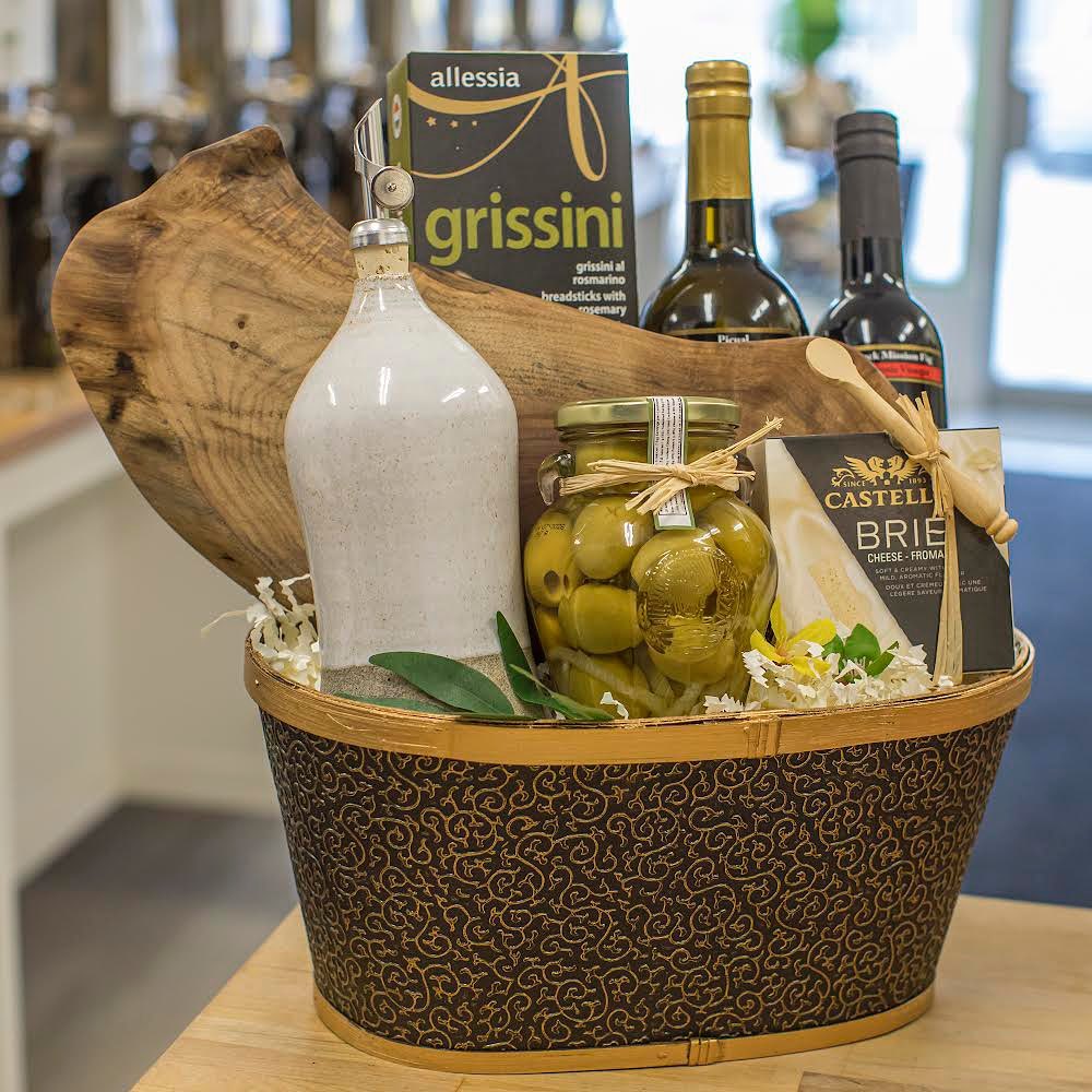 A decorative basket containing bottles of wine, a ceramic bottle, extra virgin olive oil, a jar of green olives, a package of grissini, and a box of Castello Brie cheese.